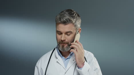 Handsome-Caucasian-man-doctor-talking-cheerfully-on-mobile-phone-and-laughing-on-the-gray-wall-background.-Close-up.